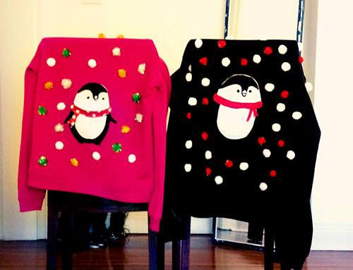 A pair of hand made christmas sweaters. One in pink, the other in black,
							both feature a penguin in felt sewed onto the front of each jumper, surrounded by many multi-coloured pompoms